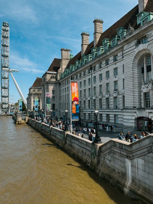 A wide view of the historic County Hall building along the River Thames in London, featuring its classic white stone facade with large arched windows, multiple chimney stacks, and a series of teal-col