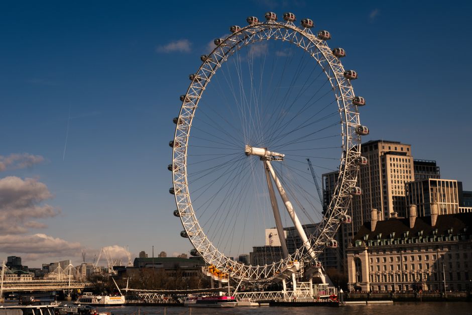 A wide view of the historic County Hall building along the River Thames in London, featuring its classic white stone facade with large arched windows, multiple chimney stacks, and a series of teal-col
